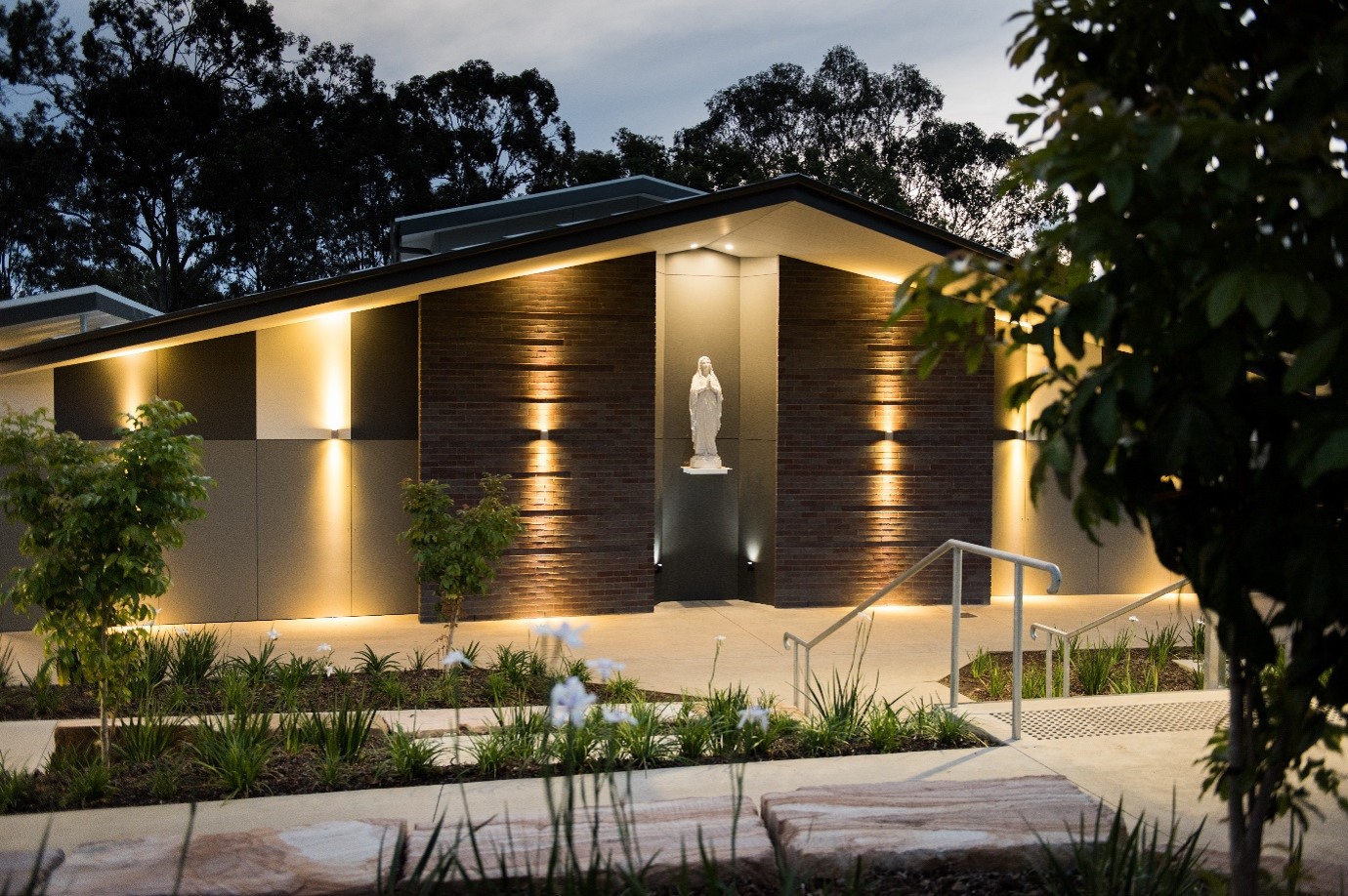 Statue of Mary located outdoors in a modern landscaped garden under lights at twilight 